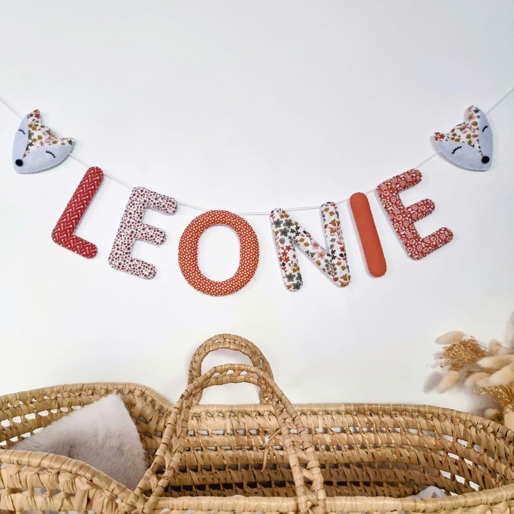 Guirlande prénom en tissu terracotta fleuri avec embouts renard, exemple « Léonie », au-dessus d'un couffin en osier