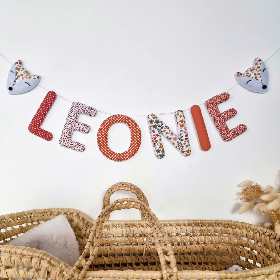 Guirlande prénom en tissu terracotta fleuri avec embouts renard, exemple « Léonie », au-dessus d'un couffin en osier