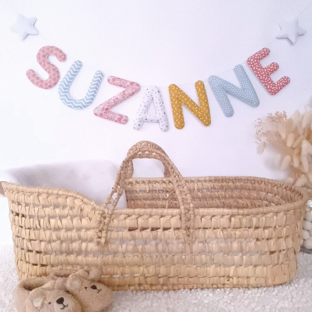 Guirlande prénom en tissu pastel multicolore avec étoiles, exemple « Suzanne », au-dessus d'un couffin en osier