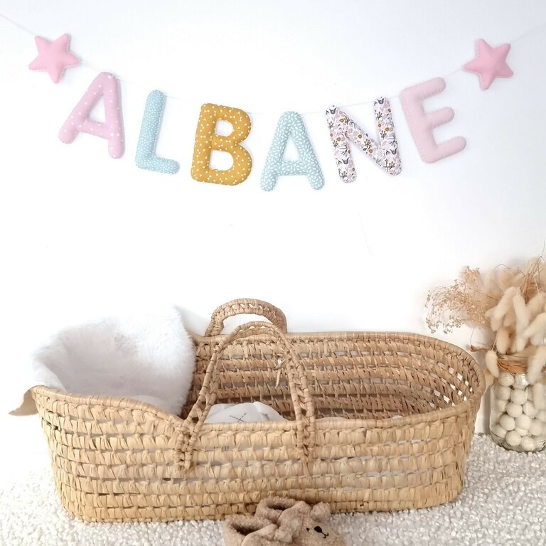 Guirlande prénom en tissu pastel rose, menthe et moutarde avec étoiles, exemple « Albane », au-dessus d'un couffin en osier